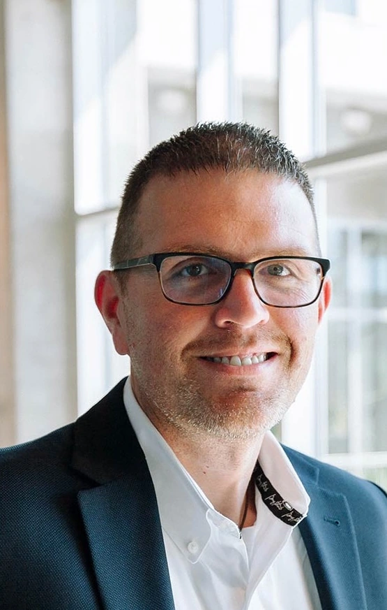 Phoenix Leader - COO Rick Childress: A smiling middle-aged man wearing a white collared shirt, a navy blue blazer and black rimmed glasses.